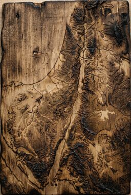  A tactile map burned into wood — rivers traced by fine scorched lines, valleys shaded by light scorch marks — mountains rising from rough, blackened texture — ancient and handmade.