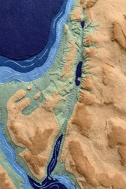  A gentle, cozy map made from layered felt — tan deserts, sage green valleys, indigo rivers — each landform a raised, plush tactile zone stitched lightly into the land's outline.